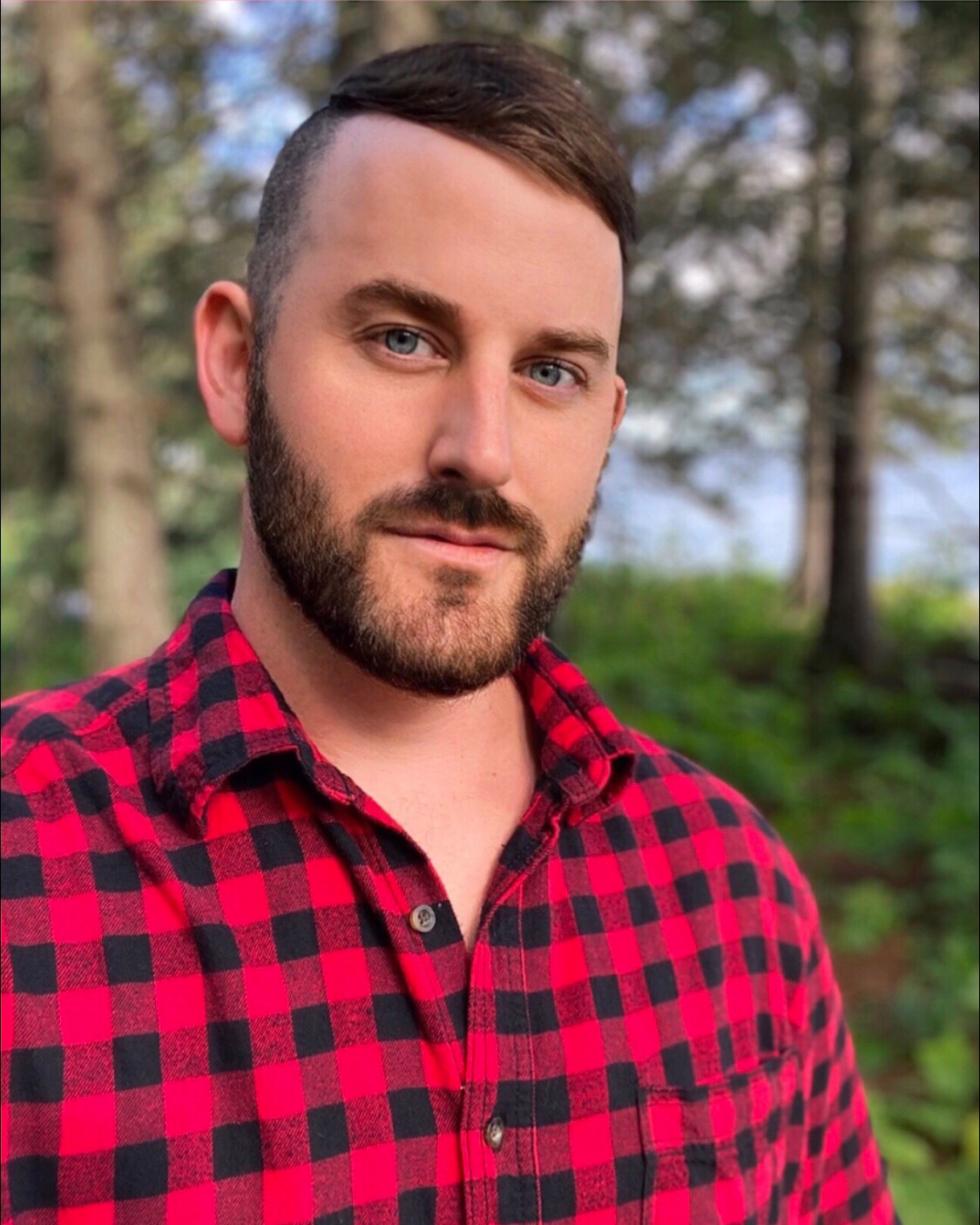Jonathan has a short beard and connecting mustache. His hair is brown. He is wearing a red and navy blue flannel. His selfie was taken in the woods.
