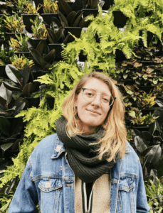 Emily Holland, a white poet with blond hair wearing a dark green scarf and jean jacket, stands in front of a lush green plant wall.
